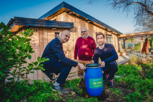 Three men with a blue plastic barrel in front of a single storey wooden building