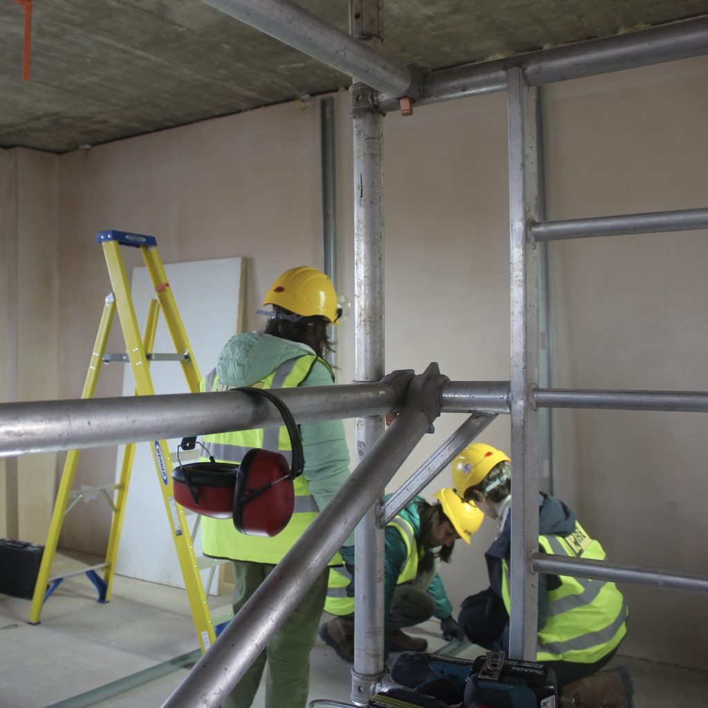 Photo shows three women in hard hats and hi-vis in a flat with bare plaster walls. They are positioning metal studs.